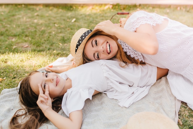 Schattige brunette meisje poseren met vredesteken op het gras terwijl haar charmante moeder strooien hoed te houden en lachen. Mooie jonge vrouw in kanten kledij gek rond met dochter op deken in park.