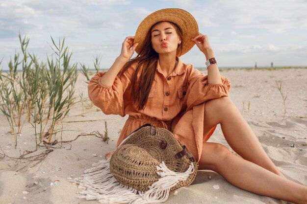 Schattige brunette meisje stuur kus en ontspannen op het strand. Modieuze linnen zomerkleding dragen. Strohoed en boho-tas.