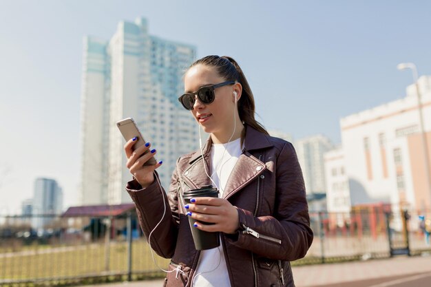 Schattige brunette vrouw in bordeaux jasje en witte tshirt messaging op telefoon buiten europese dame glimlacht oprecht en loopt buiten in zonnige stad