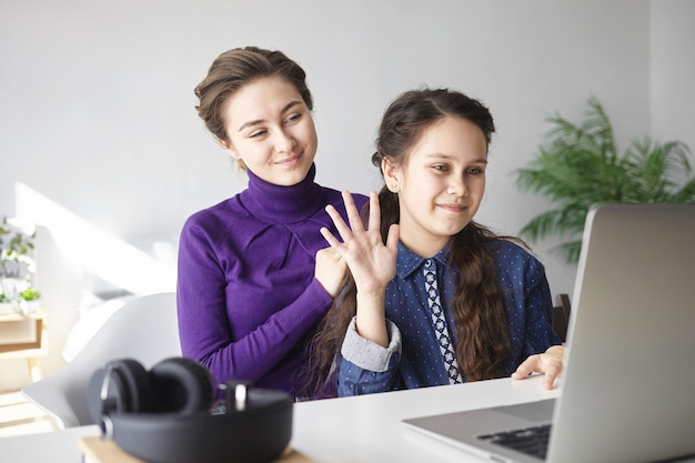 Schattige brunette vrouwelijke kind zwaaiende hand op laptop scherm met behulp van videochat samen met haar jonge moeder in de slaapkamer