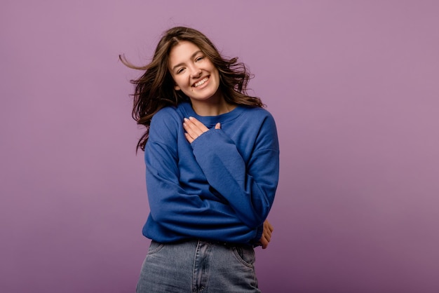 Schattige jonge blanke brunette vrouw in blauwe hoodie glimlachend terwijl ze naar de camera kijkt op paarse achtergrond Mensen emoties levensstijl concept