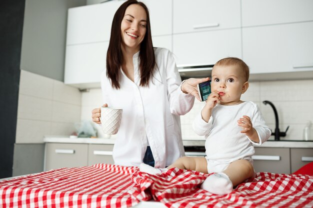 Schattige jonge moeder die telefoon geeft aan baby en glimlacht