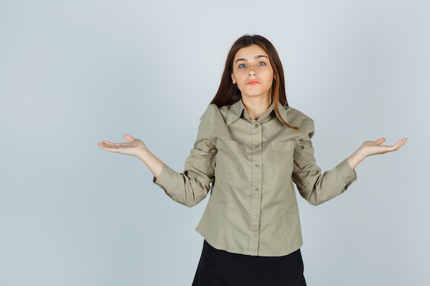 Schattige jonge vrouw die hulpeloos gebaar toont door haar schouders op te halen in shirt, rok en verward, vooraanzicht.