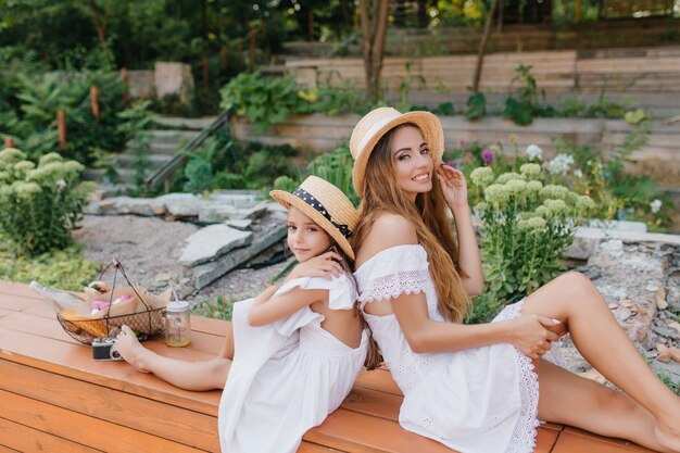 Schattige jonge vrouw in goed humeur genieten in prachtig park met stenen en bloemen. Outdoor Portret van een klein meisje in jurk met open rug zitten in de buurt van moeder in trendy schipper.