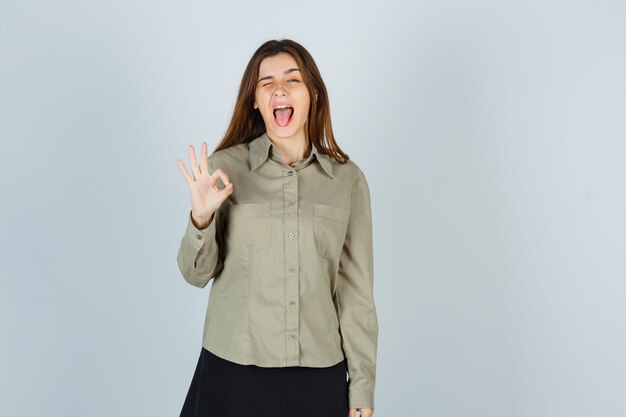 Schattige jonge vrouw in shirt, rok die een goed gebaar toont terwijl ze knippert, tong uitsteekt en er gek uitziet, vooraanzicht.