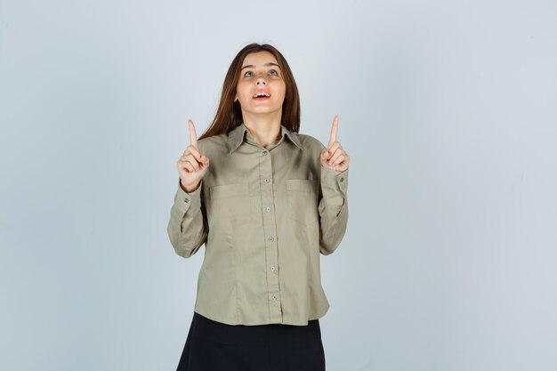 Schattige jonge vrouw in shirt, rok die omhoog wijst en er hoopvol uitziet, vooraanzicht.