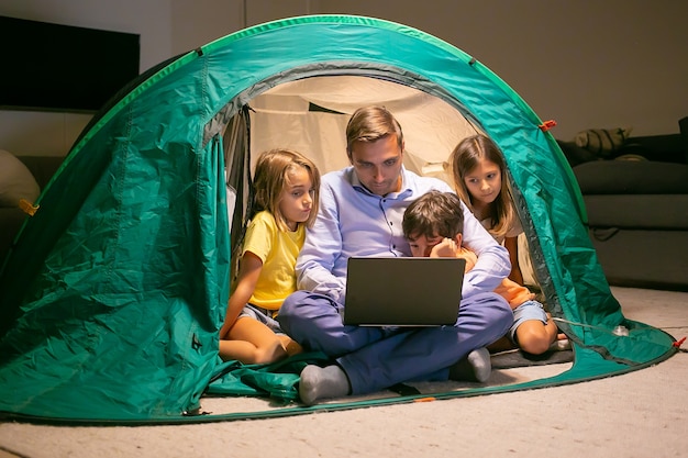 Schattige kinderen ontspannen met vader in tent thuis en kijken naar film op laptopcomputer. gelukkige kinderen en liefdevolle vader zitten in tent met licht. jeugd, familie tijd en weekend concept