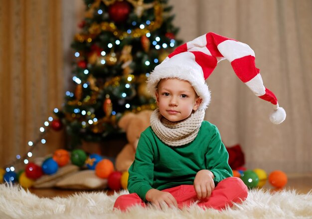 Schattige kleine jongen in kerstmuts met kerstboom op de achtergrond.