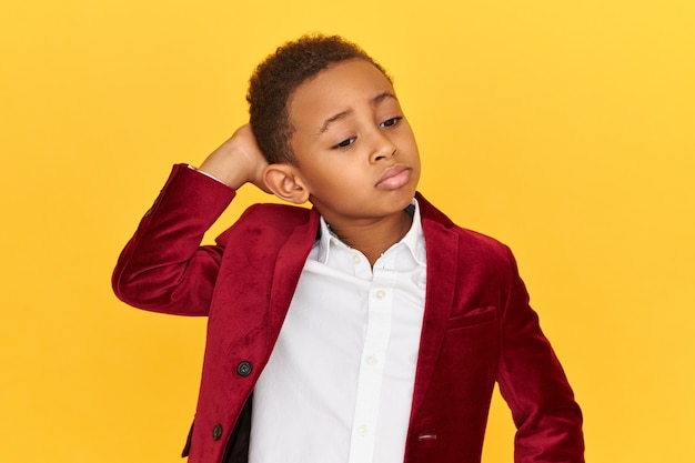 Schattige kleine jongen met donkere huid die lippen pruilt en de hand op de achterkant van zijn nek houdt, verwarde gezichtsuitdrukking gefrustreerd heeft, geïsoleerd poseren.