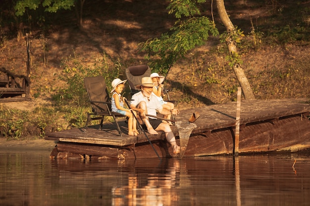 Schattige kleine meisjes en hun opa zijn aan het vissen in het meer of de rivier. Rusten op pier in de buurt van water en bos in de tijd van de zonsondergang van zomerdag. Concept van familie, recreatie, kindertijd, natuur.