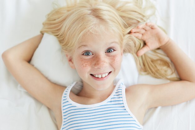 Schattige kleine vrouwelijke kind met blond haar, blauwe ogen en sproeten gezicht, lacht vrolijk terwijl ontspannen op bed, liggend op wit kussen.