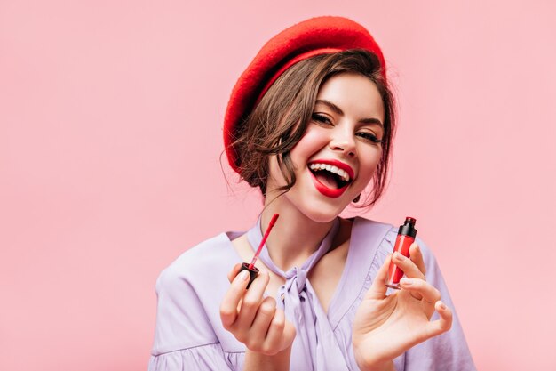 Schattige krullende vrouw schildert haar lippen met rode lippenstift en lacht op roze achtergrond.