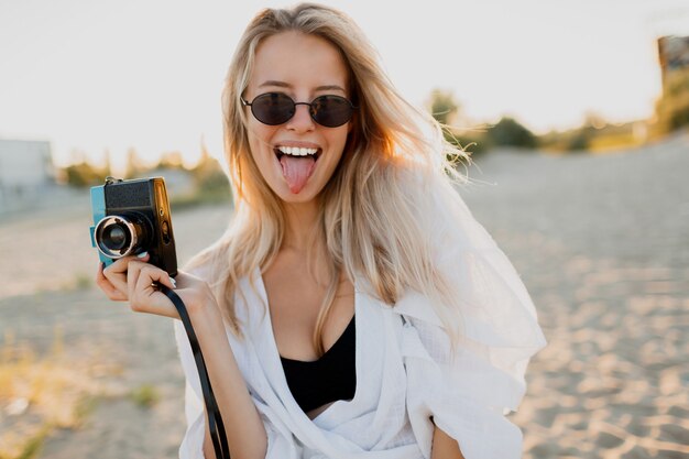 Schattige meid met retro camera grimassen maken en poseren op het strand in de buurt van de oceaan. Zomervakantie. Prachtig zonlicht.