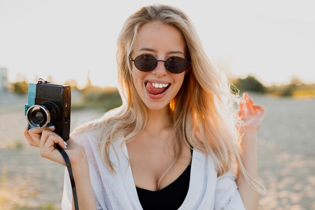 Schattige meid met retro camera grimassen maken en poseren op het strand in de buurt van de oceaan. Zomervakantie. Prachtig zonlicht.