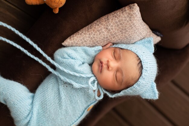 Schattige pasgeboren mooie en sympathieke babyjongen die op bruine bank en hoofdkussen in blauwe gehaakte pijamas slaapt