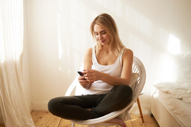 Schattige tiener meisje zit in comfortabele fauteuil in haar slaapkamer bedrijf mobiele telefoon, vrienden online messaging, plannen maken voor weekend. Schattige jonge vrouw die op internet surft via mobiel met behulp van wifi