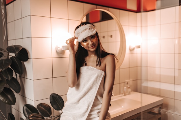 Schattige vrouw in handdoek en gouden patches onder ogen poseren in lichte badkamer