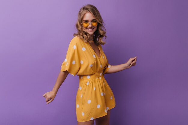 Schattige vrouw in vintage oranje kleding lachen naar de camera. Portret van spectaculair meisje met golvend blond meisje geïsoleerd op paars.