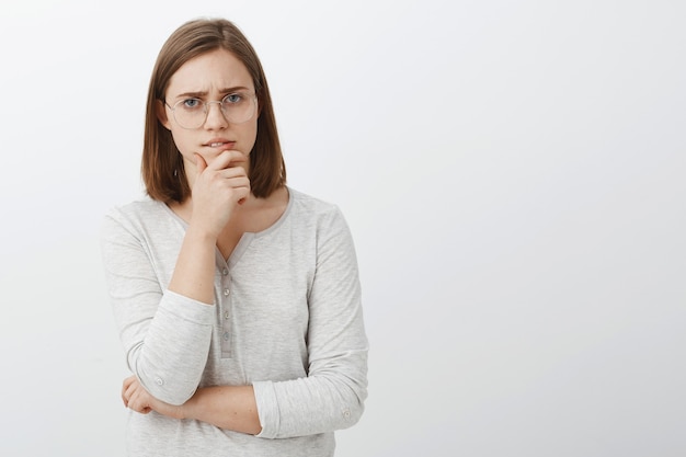 Schattige vrouwelijke geek probeert hard wiskunde raadsel op te lossen, nadenkend over de witte muur, kin wrijven tijdens brainstorm, kijken tijdens het nemen van beslissingen of denken poseren over grijze muur