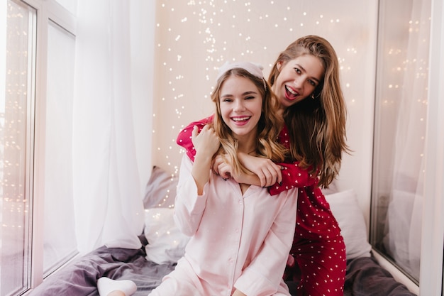 Schattige zusjes dragen trendy pyjama's die in de slaapkamer voor de gek houden. Verbazingwekkende blanke meisjes die in de ochtend energie uitdrukken.