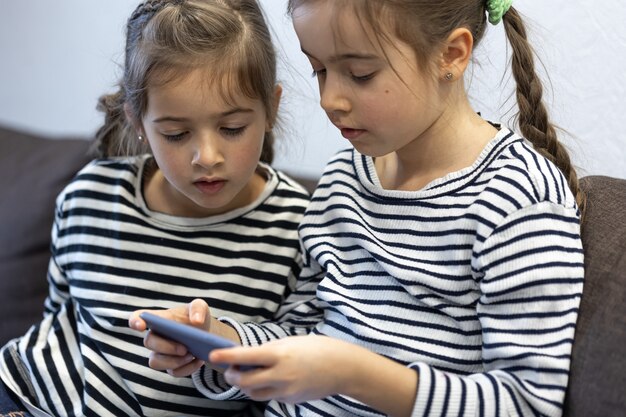 Schattige zusjes gebruiken telefoons terwijl ze thuis op de bank zitten.