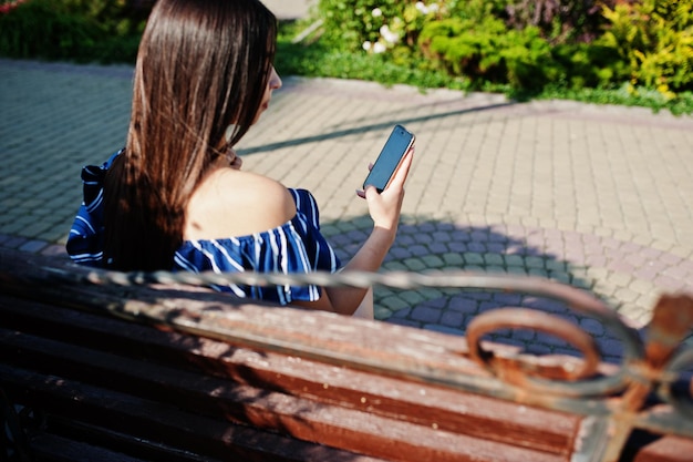 Schitterend brunette meisje zittend op een bankje in de straat van de stad en kijkend naar telefoonslijtage op een blauw gestreepte jurk