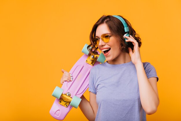 Schitterend meisje in geel-gekleurde zonnebril luisteren muziek in hoofdtelefoons. Portret van modieus vrouwelijk model met skateboard dat favoriete liedje zingt.