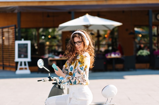 Schitterend meisje met lang krullend haar shirt met bloemmotief dragen, zittend op een scooter en met mobiele telefoon