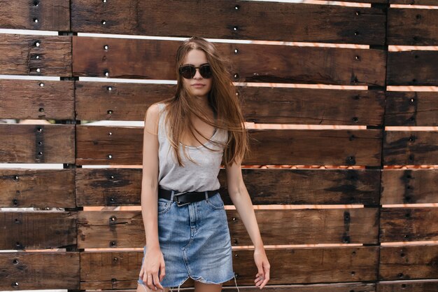 Schitterend wit meisje in vintage denim rok poseren op houten muur. Buiten schot van ernstige langharige jonge vrouw.