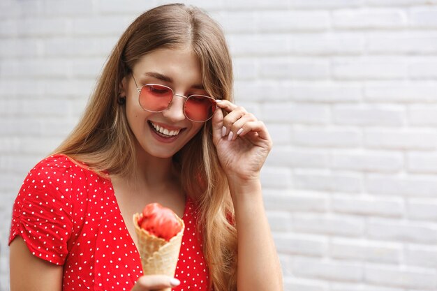 Schitterende vrouw die in zomerjurk en zonnebril staat en ijs eet, glimlacht en ziet er gelukkig uit Vrouwelijke reiziger die buiten op vakantie loopt
