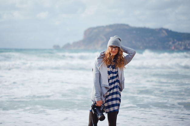 Schitterende vrouw in grijze hoodie en hoeden die camerand glimlachen die in strand overdag met overzees glimlachen