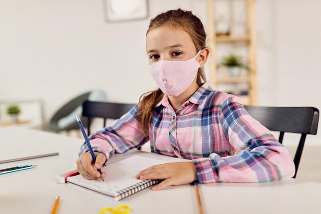 Schoolmeisje met beschermend gezichtsmasker dat in notitieboekje schrijft tijdens thuisonderwijs