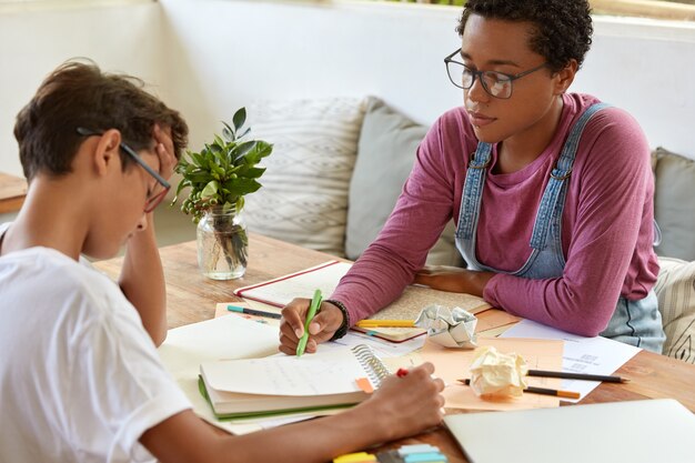 Schoolonderwijs en thuisbegeleiding concept. Horizontale opname van zwarte slimme Afrikaanse Amerikaanse vrouw beantwoordt een vraag van een schooljongen die hoofdpijn heeft en flip-over of diagram niet kan begrijpen