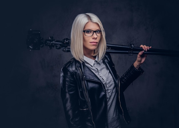 Schoonheid blonde vrouwelijke fotograaf in bril met een witte blouse en zwart leren jas heeft een professionele camera met een statief, poseren in een studio. geïsoleerd op een donkere achtergrond.