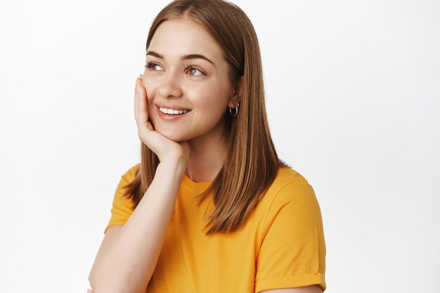 Schoonheid. Close-up portret van dromerige gelukkige vrouw, glimlachend opzij kijkend met doordacht dagdromen gezicht, schone gloeiende huid aan te raken, staande op een witte achtergrond.