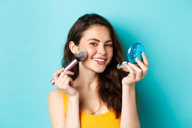 Schoonheid. Close-up van een aantrekkelijke vrouw die in de spiegel kijkt en een penseel gebruikt om make-up aan te brengen, tevreden glimlachend in de camera, staande tegen een blauwe achtergrond.