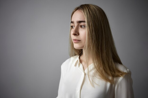 Schoonheid en elegantie concept. Prachtige mooie jonge blonde vrouw in zijdeachtig shirt met bovenste botton ongedaan gemaakt poseren geïsoleerd tegen lege grijze studio muur achtergrond met copyspace voor uw advertentie