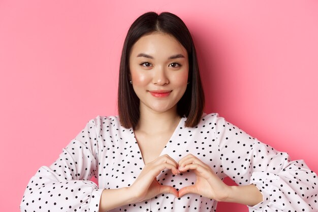 Schoonheid en lifestyle concept. Close-up van een mooie aziatische vrouw die een hartteken toont, glimlacht en romantisch voelt op Valentijnsdag, staande over een roze achtergrond.