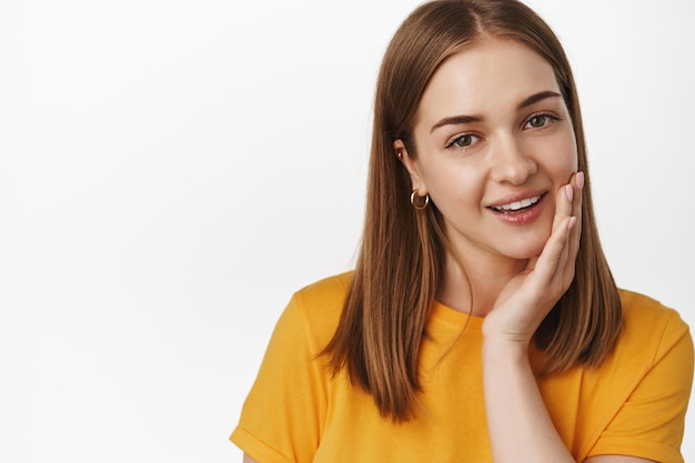 Schoonheid en vrouwen. Close up van aantrekkelijk blond meisje touch schoon, geen make-up gloeiende huid, hand op de wang vasthouden, concept van huidverzorging en gezichtscosmetica, witte achtergrond.