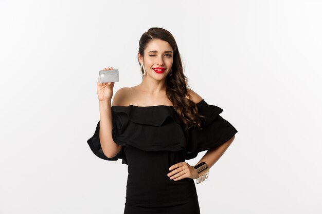 Schoonheid en winkelconcept. Modieuze vrouw in zwarte kleding, die make-up draagt, knipoogt naar de camera en creditcard toont, die zich over witte achtergrond bevindt.