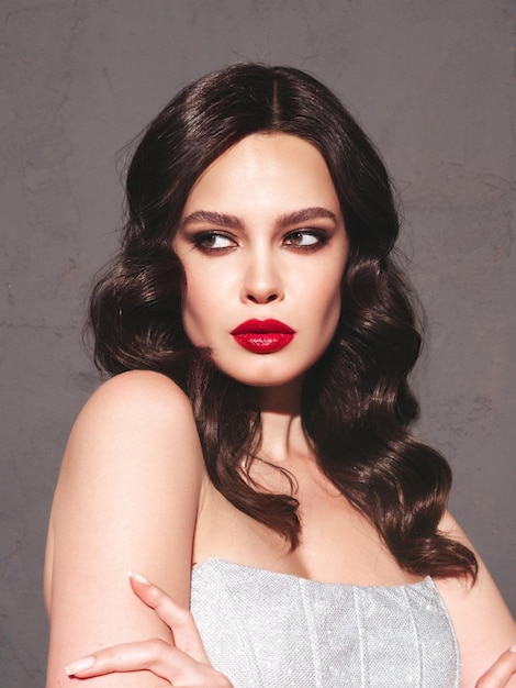 Schoonheid portret van jonge brunette vrouw met avond make-up en perfecte schone huid Sexy model met krullend haar poseren in studio in de buurt van donkere muur met rode heldere natuurlijke lippen In zilveren jurk