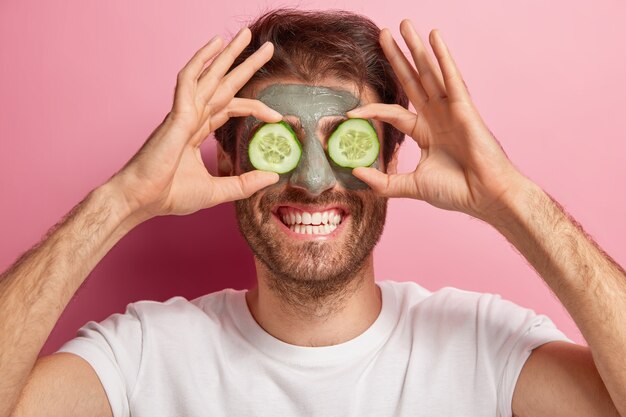 Schoonheid portret van vrolijke man vormt met kleimasker op gezicht, twee plakjes komkommer op de ogen, draagt een wit t-shirt, heeft brede glimlach en borstelharen