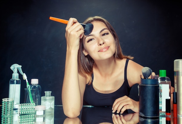 Schoonheid vrouw make-up toe te passen. Mooi meisje in de spiegel kijken en cosmetica met een grote borstel toe te passen. Ochtend, make-up en menselijke emoties concept. Kaukasisch model in de studio