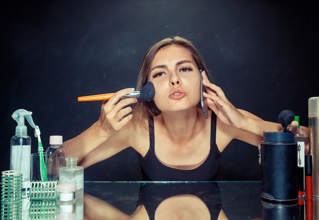 Schoonheid vrouw make-up toe te passen. Mooi meisje in de spiegel kijken en cosmetica met een grote borstel toe te passen. Ochtend, make-up en menselijke emoties concept. Kaukasisch model in de studio