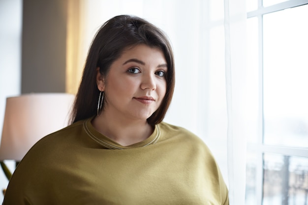 Schoonheid, vrouwelijkheid, stijl en mode-concept. Portret van mooie overgewicht jonge donkerharige Europese vrouw met nette make-up poseren in een stijlvolle hotelkamer interieur bij raam en glimlachen