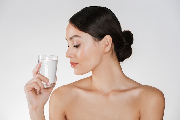 Schoonheidsportret van jonge gelukkige vrouw met haar in broodje die transparant glas van nog water bekijken die ter beschikking houden, dat over wit wordt geïsoleerd