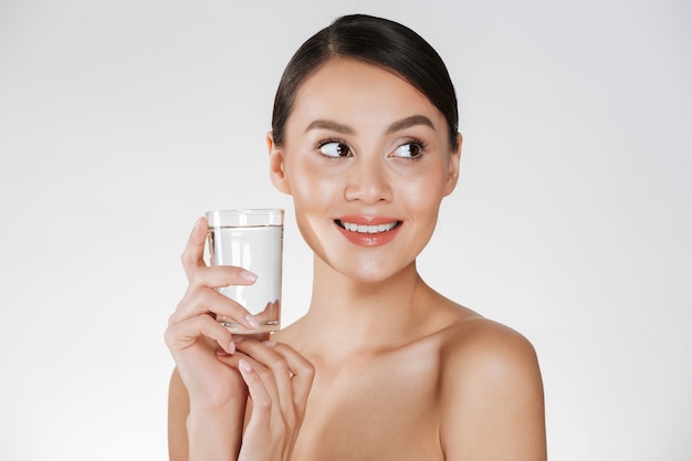 Schoonheidsportret van jonge gelukkige vrouw met haar in broodje het drinken nog water van transparant glas, dat over wit wordt geïsoleerd