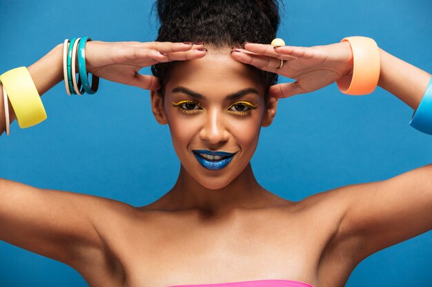 Schoonheidsportret van schitterende afro Amerikaanse vrouw met maniermake-up en armbanden op handen die op camera met glimlach stellen geïsoleerd, over blauwe muur