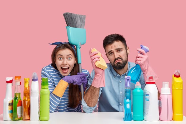 Schoonmaak concept. Trieste jongeman houdt reinigingsmiddel en spons vast, heeft een ongelukkige uitdrukking, beu met spiegel wassen, vrolijke dame wijst naar man, draagt borstel