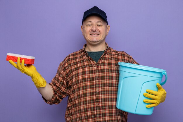 Schoonmaak man in geruit hemd en pet met rubberen handschoenen met spons en emmer kijkend naar camera gelukkig en positief glimlachend vrolijk staande over paarse achtergrond